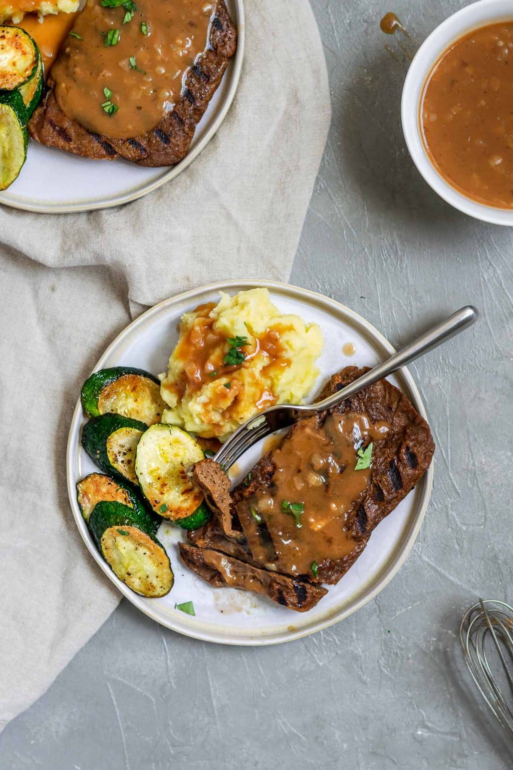 Gegrillte hausgemachte Seitan-Steaks mit Kartoffelpüree und Schalottensoße mit einer Beilage aus Zucchini und einer Garnierung aus Petersilie. Das Steak wird mit einem Biss auf einer Gabel geschnitten und es wird extra Soße auf der Seite serviert.